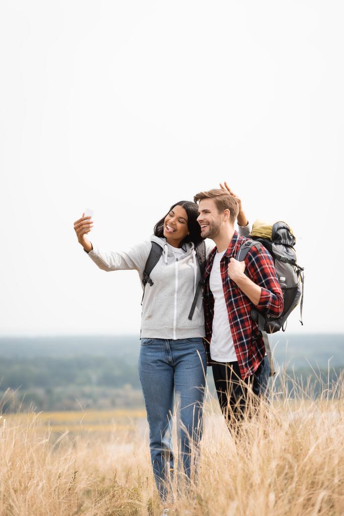 Uśmiechnięci wielonarodowi podróżnicy robiący selfie ze smartfonem na łące  - Zdjęcie, obraz