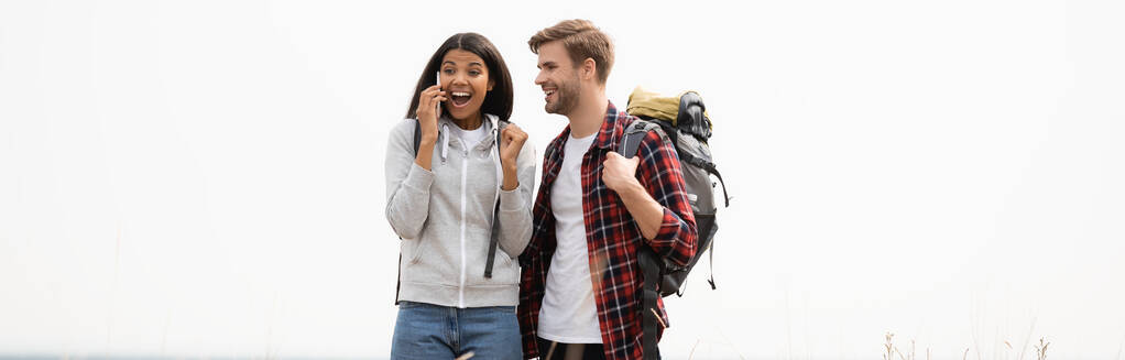 Cheerful african american woman talking on smartphone near boyfriend with backpack isolated on grey, banner  - Photo, Image