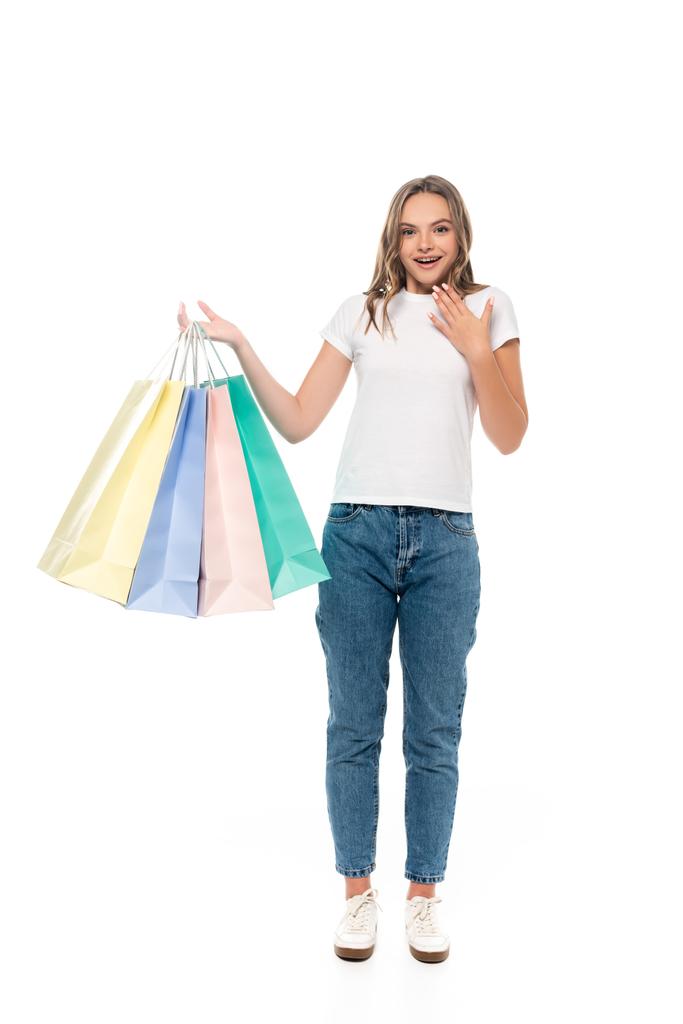 mujer joven emocionada sosteniendo coloridas bolsas de compras y mirando a la cámara aislada en blanco - Foto, imagen