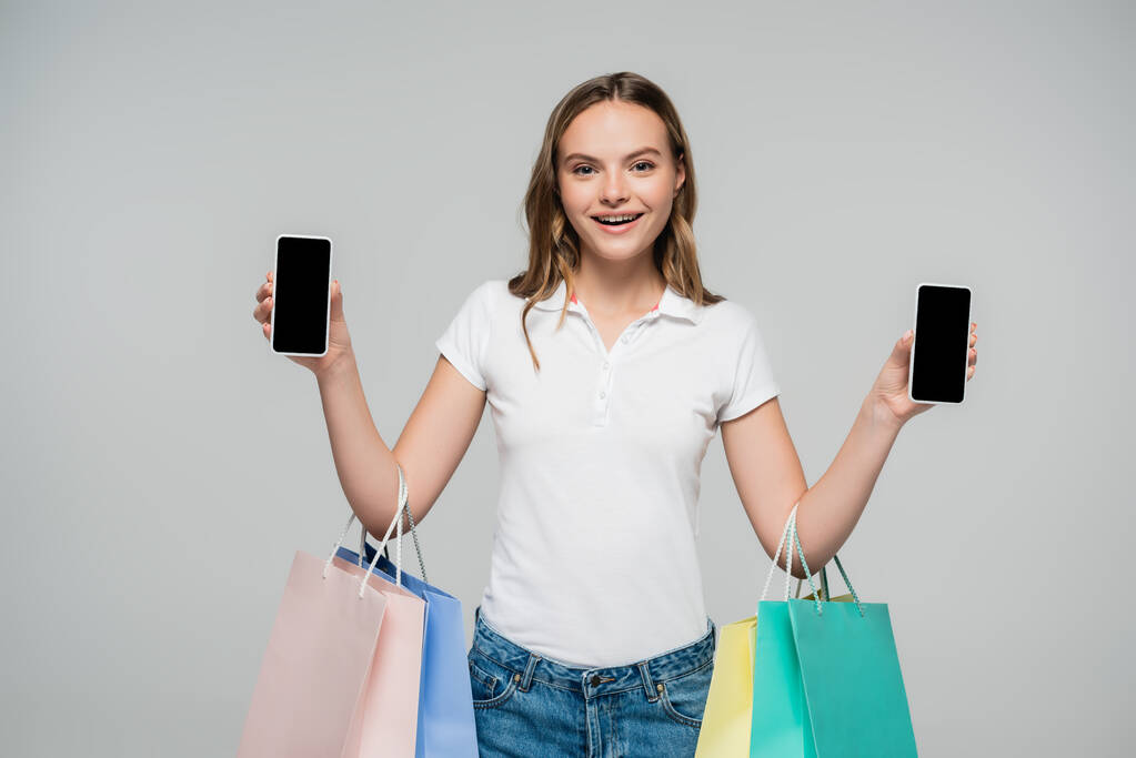 mujer alegre sosteniendo teléfonos inteligentes con pantalla en blanco y bolsas de compras aisladas en gris, concepto de viernes negro  - Foto, Imagen