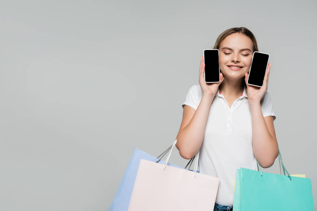 mujer complacida con los ojos cerrados sosteniendo teléfonos móviles con pantalla en blanco y bolsas de compras aisladas en gris, concepto de viernes negro  - Foto, imagen