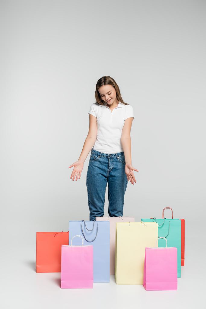 mujer alegre de pie y señalando con las manos a las bolsas de compras en gris, concepto de viernes negro  - Foto, imagen