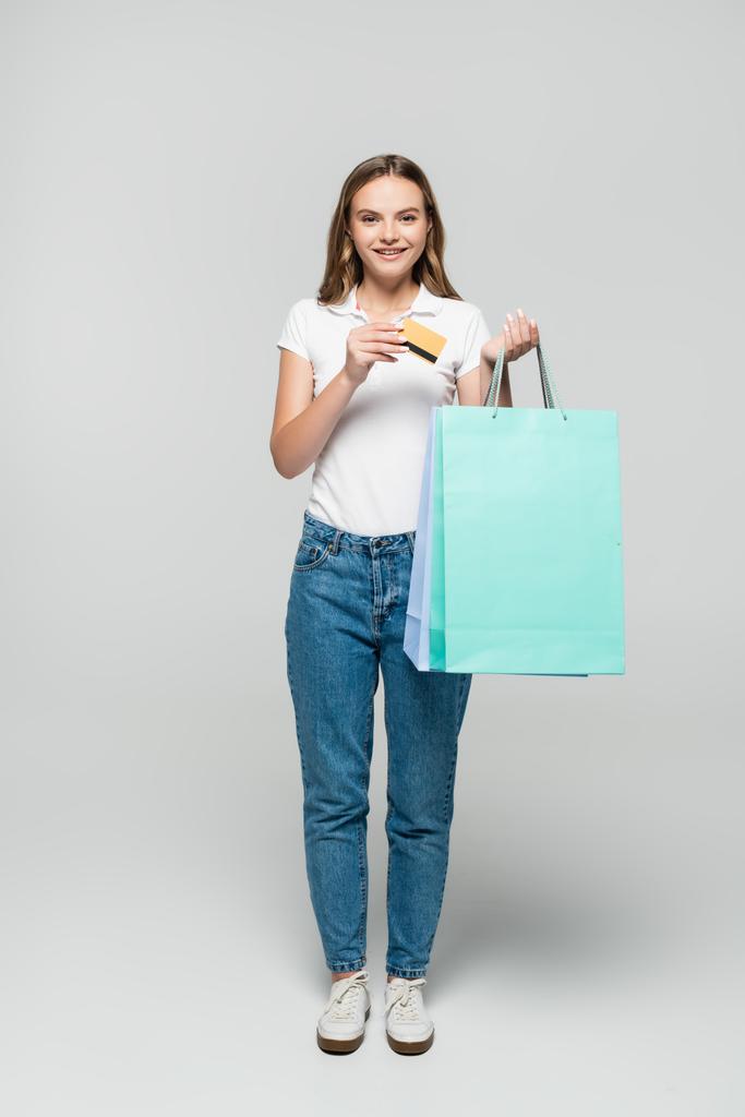 alegre joven con tarjeta de crédito y bolsas de compras azules en gris, concepto de viernes negro - Foto, imagen