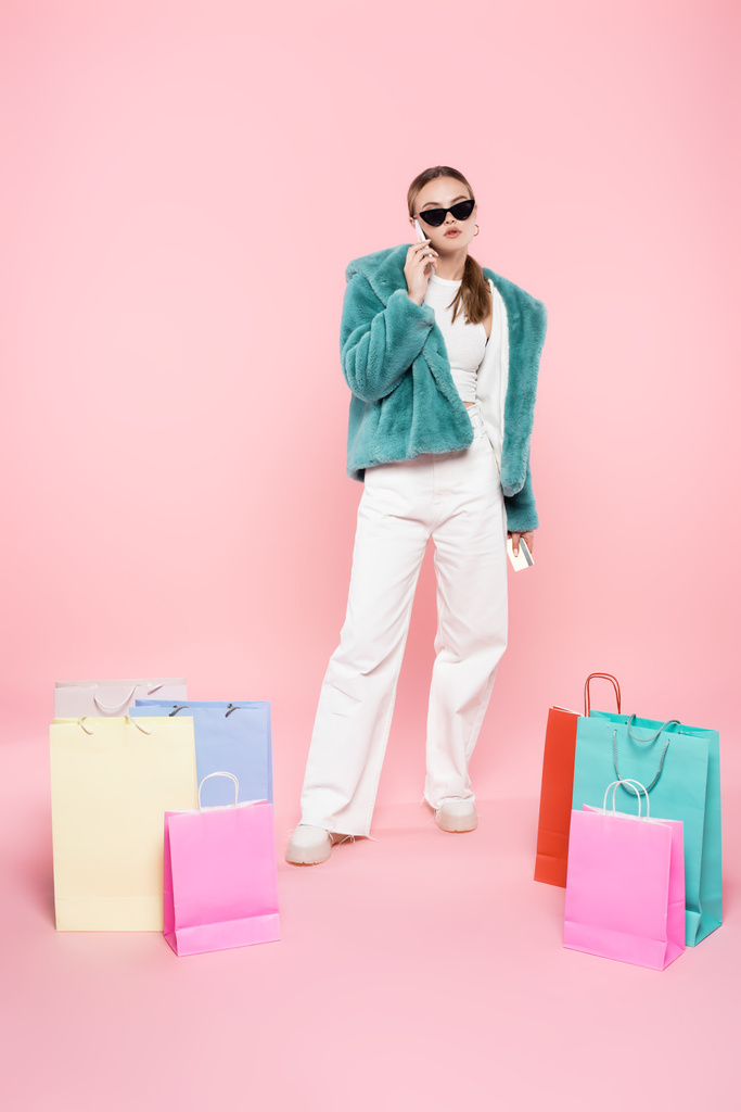 stylische Frau mit Sonnenbrille, die am rosa, schwarzen Freitag neben Einkaufstüten auf dem Smartphone spricht  - Foto, Bild