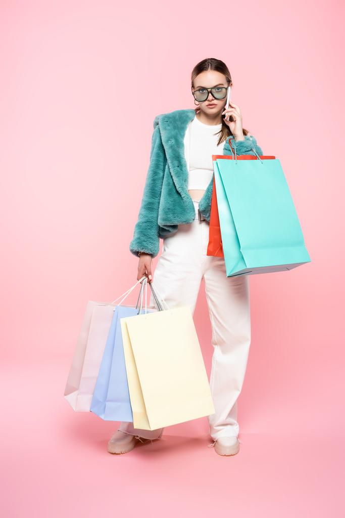 mujer de moda en gafas de sol hablando en el teléfono inteligente cerca de bolsas de compras en rosa, concepto de viernes negro  - Foto, imagen