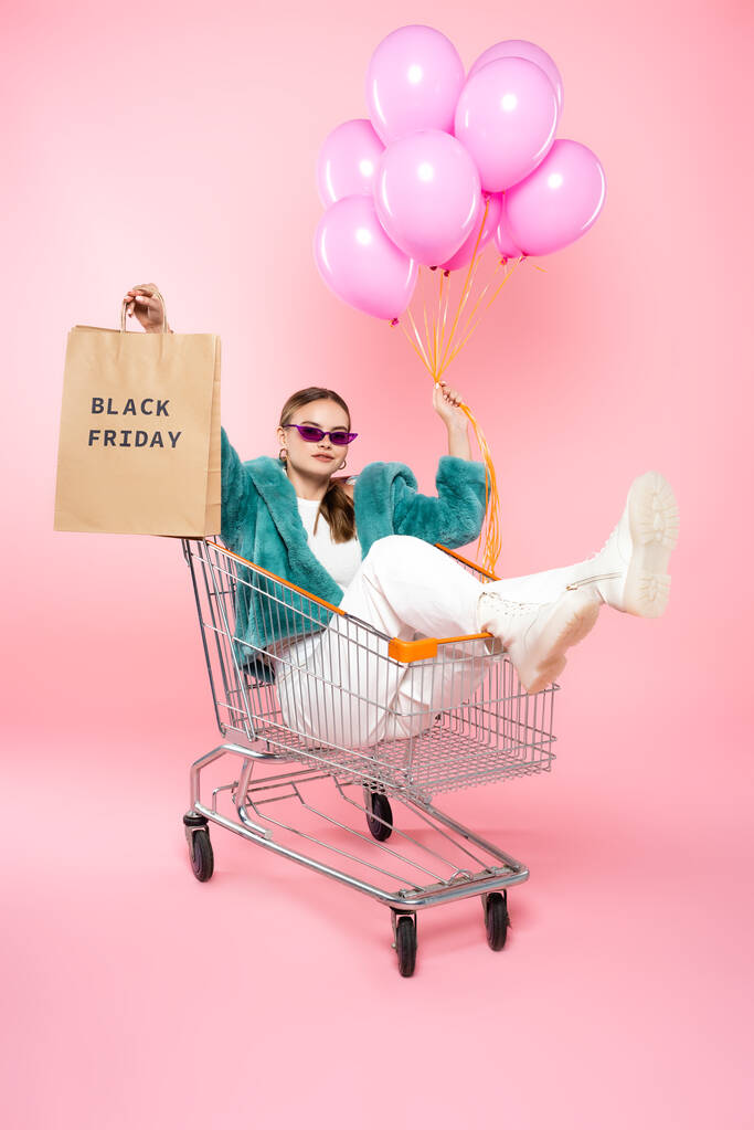 стильна жінка в сонцезахисних окулярах, що сидить у візку з чорною п'ятничною пляшкою на торговому мішку і тримає повітряні кулі на рожевому
  - Фото, зображення