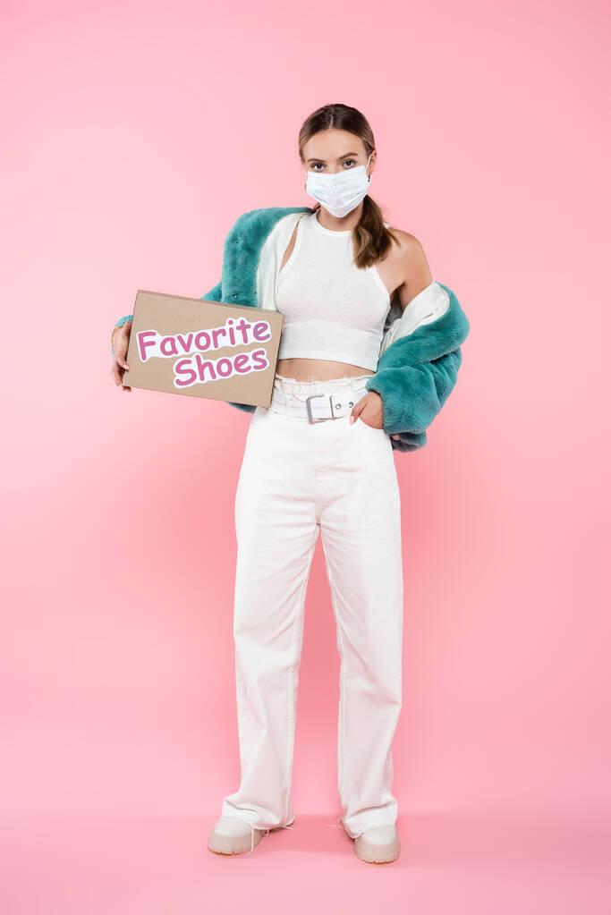 jeune femme dans le masque médical tenant boîte avec des chaussures préférées lettrage tout en se tenant avec la main dans la poche sur rose - Photo, image