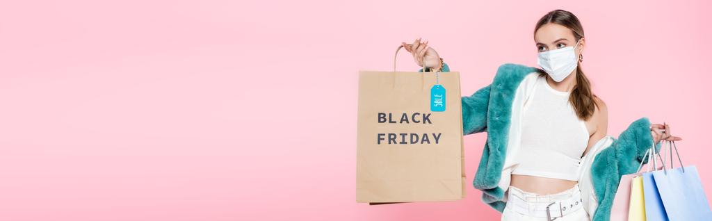 plano panorámico de cliente joven en máscara médica sosteniendo bolsa de compras con letras de viernes negro y etiqueta de venta aislado en rosa  - Foto, imagen