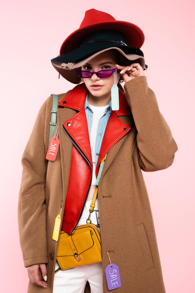 модна молода жінка в капелюхах торкається сонцезахисних окулярів з тегом продажу на рожевому
 - Фото, зображення