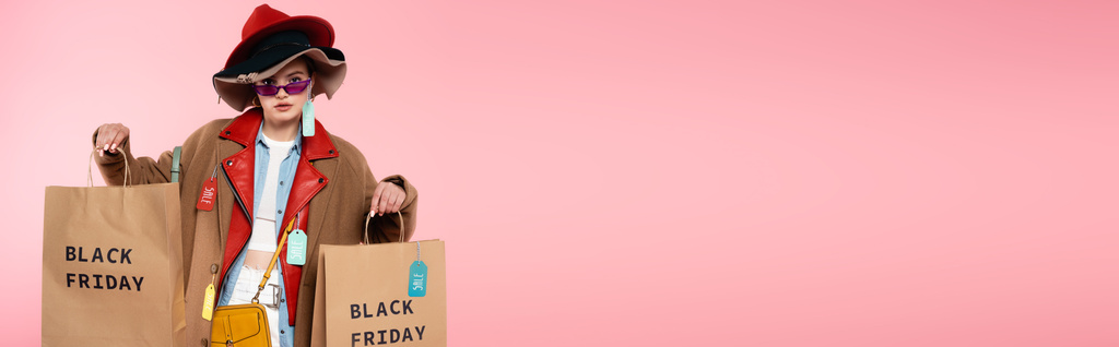 horizontales Bild einer trendigen Frau in Sonnenbrille und Hut mit Verkaufsetiketten mit Einkaufstaschen auf rosa, schwarzem Freitag-Konzept - Foto, Bild
