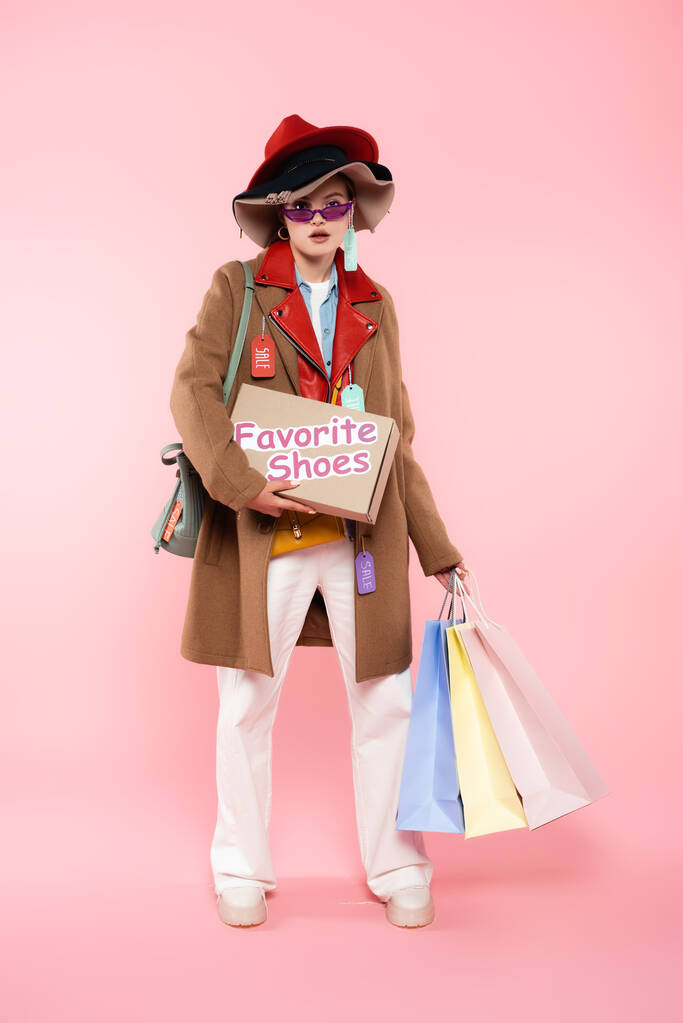 fiatal nő napszemüveg és kalap eladó címkék tartó doboz kedvenc cipő felirat és bevásárló táskák rózsaszín, fekete péntek koncepció - Fotó, kép