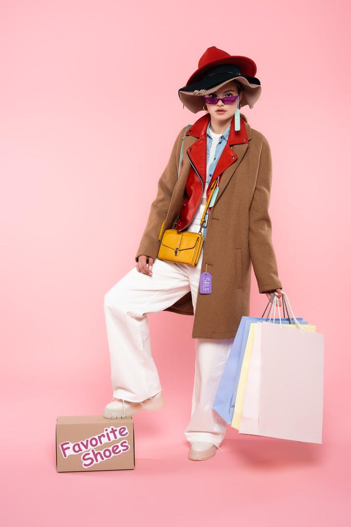 kobieta w okularach przeciwsłonecznych i kapeluszach ze sprzedażą tagów trzymających torby na zakupy i stojąca w pobliżu pudełka z ulubionymi butami na różowym - Zdjęcie, obraz