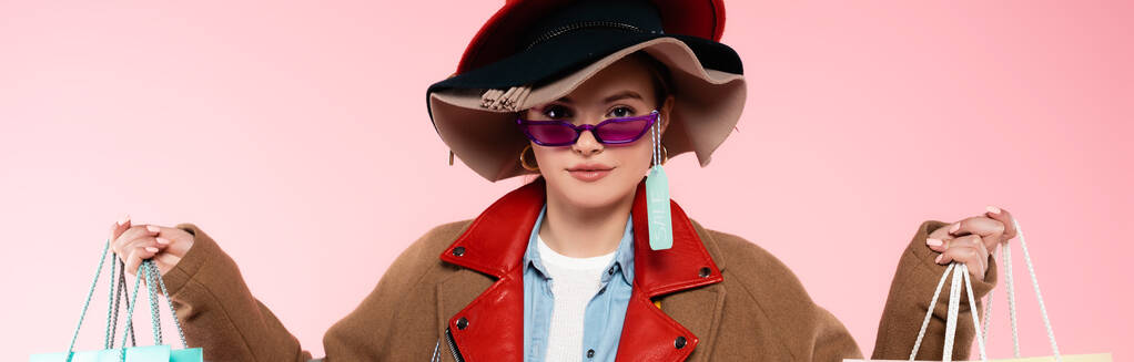 colpo panoramico di donna in occhiali da sole e cappelli con tag vendita tenendo borse della spesa isolate su rosa - Foto, immagini