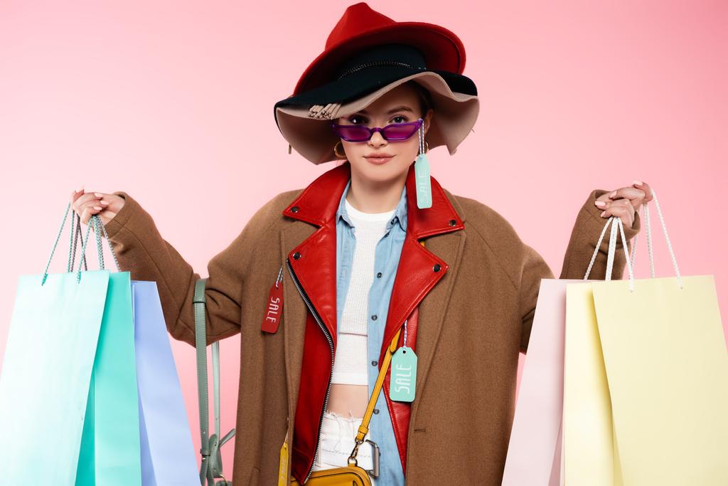 μοντέρνα γυναίκα με γυαλιά ηλίου και καπέλα με ετικέτες πώλησης κρατώντας τσάντες για ψώνια σε ροζ - Φωτογραφία, εικόνα