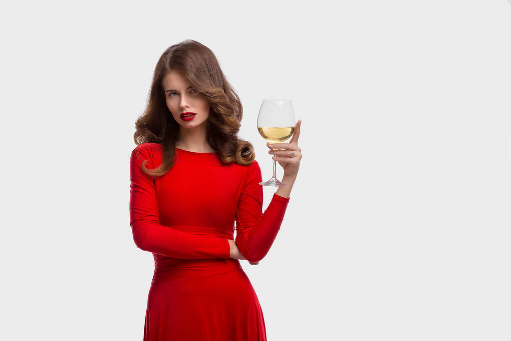 Frau mit Make-up, Frisur wehendem roten Kleid posiert mit einem Glas Wein auf weißem Hintergrund, isoliert - Foto, Bild