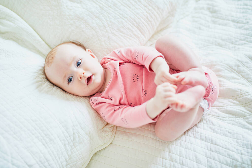 Menina deitada na cama de costas e tocando os dedos dos pés. Criança saudável feliz em roupas rosa. Criança infantil em berçário ensolarado - Foto, Imagem