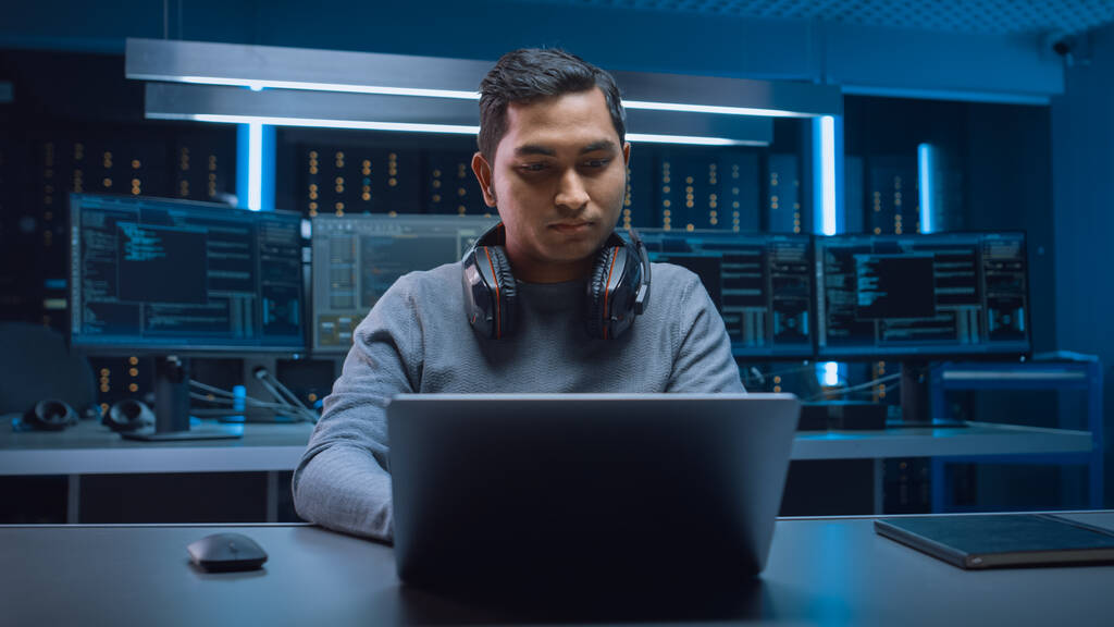 Portret van Handsome Indiase Software Developer Hacker Gamer dragen headset zitten aan zijn bureau en werken spelen op laptop. In de achtergrond Dark Neon High Tech omgeving met meerdere beeldschermen - Foto, afbeelding