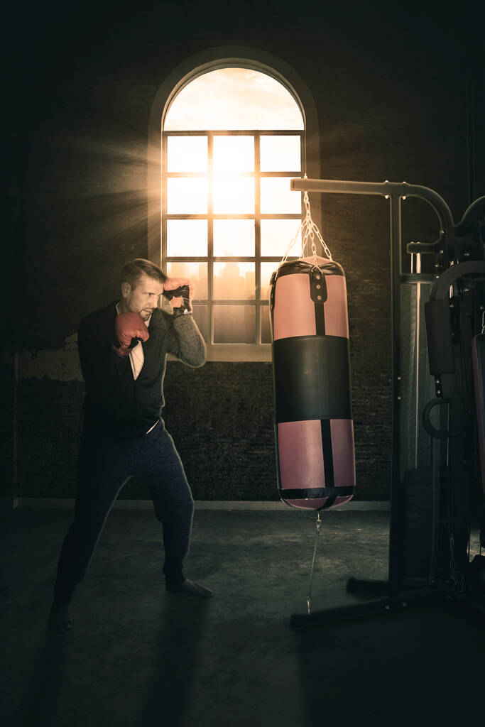 Kaukasischer Mann trainiert mit Boxsack im Fitnessstudio, während er mit Sonnenlicht durch das Fenster steht - Foto, Bild