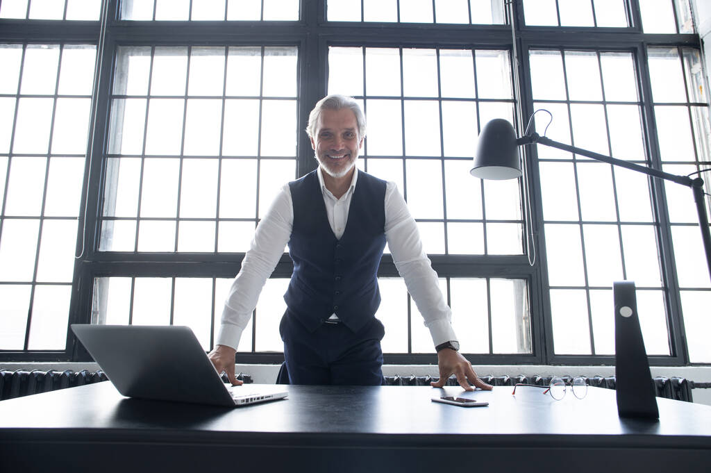 Уверенный бизнесмен, позирующий, опираясь на стол в офисе. - Фото, изображение