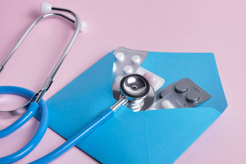 pílulas em um envelope azul e um estetoscópio médico em um fundo rosa - Foto, Imagem