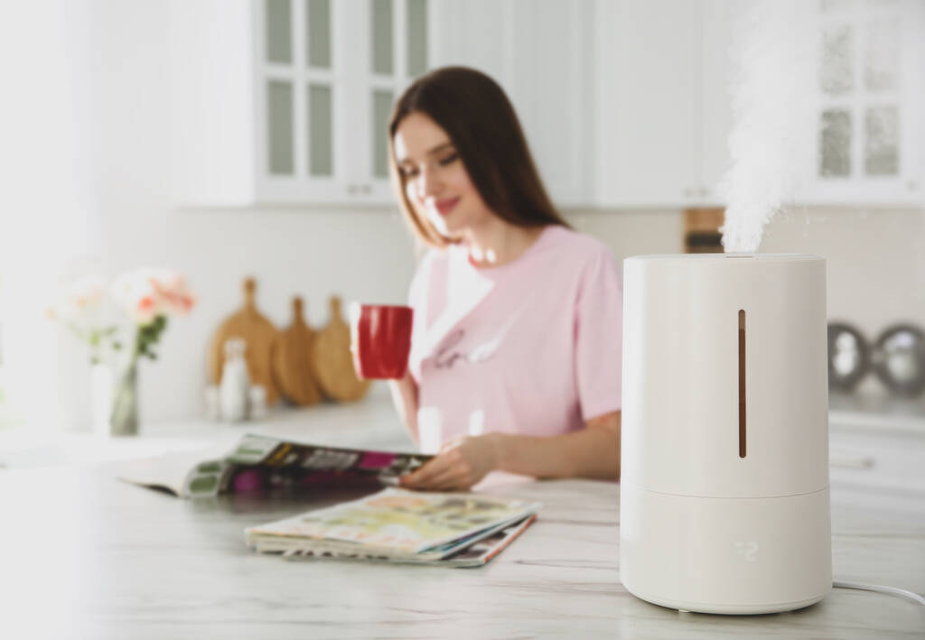 Современный увлажнитель воздуха и размытая женщина пьет кофе на кухне - Фото, изображение