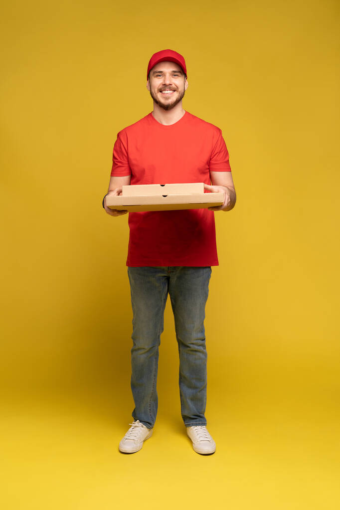 Happy man van de bezorgdienst in rood t-shirt en pet geven bestelling en houden pizza doos geïsoleerd over geel - Foto, afbeelding