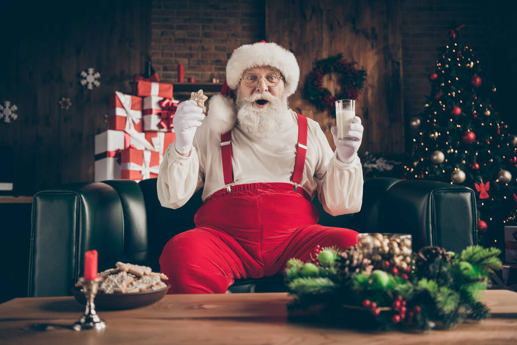 ショックを受けた灰色のひげのサンタクラスは居心地の良いソファの腕時計を座っているクリスマス映画印象的なドリンクミルクガラス食べるジンジャーブレッドクッキー家の中で赤い帽子の帽子の頭の摩耗屋内ノエルの装飾 - 写真・画像