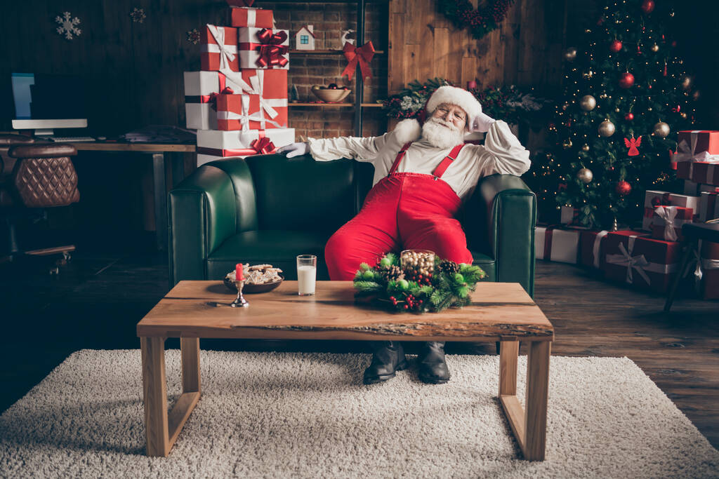 フルサイズの写真ジョリーホリーサンタクラスの座ってソファの腕時計クリスマス映画は、牛乳のガラスの生姜クッキーと居心地の良いテーブルを持っています家の中で赤い衣装を身に着けている屋内新年の装飾 - 写真・画像