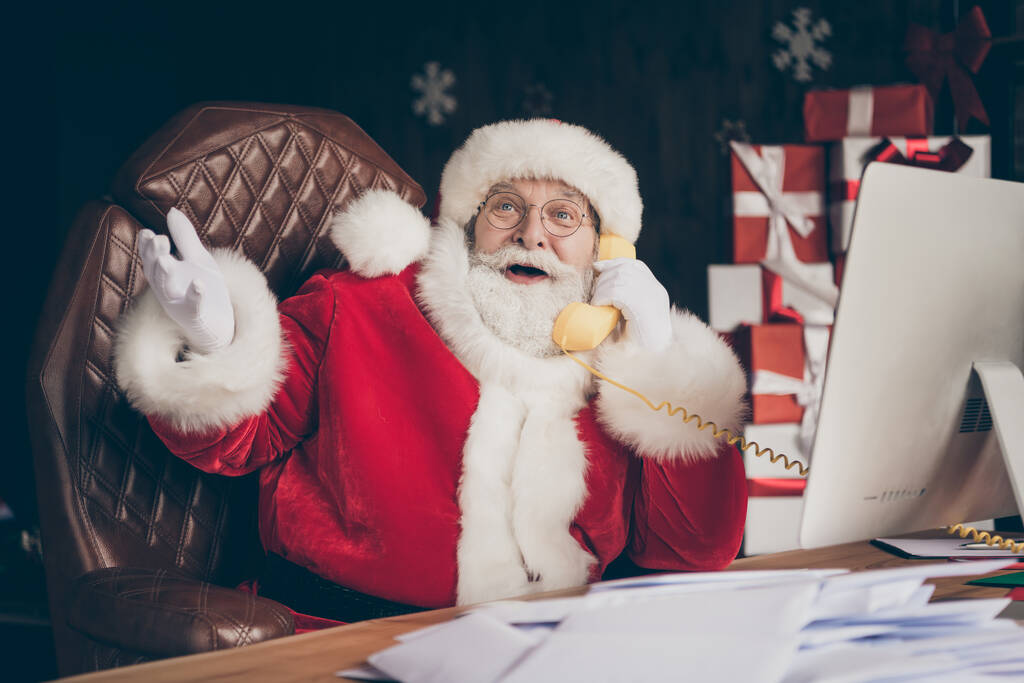 Kuva hauska harmaa parta Joulupukki istua pöytä puhelu puhua puhelin on x-mas joululahja läsnä toimitus sopimus pukeutua punainen lippis talossa sisätiloissa - Valokuva, kuva