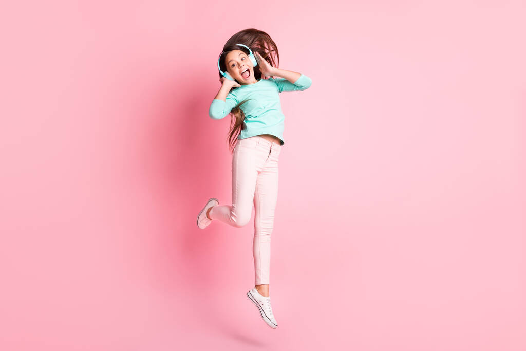Фотография полного размера тела симпатичной милой маленькой и сумасшедшей латиноамериканской леди, прыгающей с открытым ртом, руки-уши в наушниках бирюзовые брюки белого цвета на розовом фоне - Фото, изображение