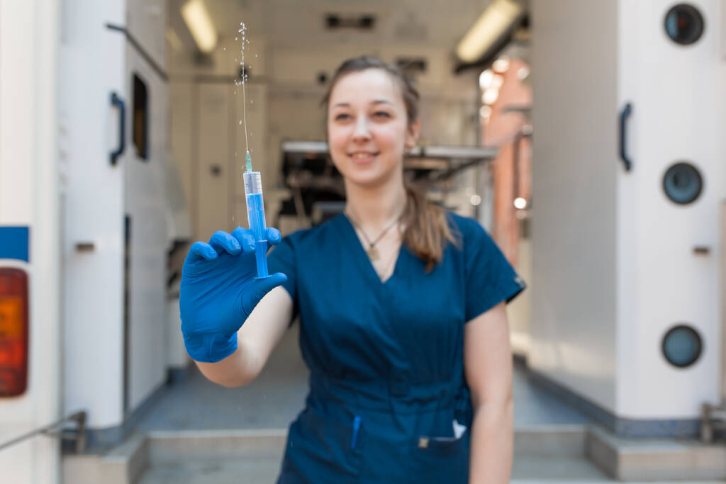 Jovem enfermeira profissional em uniforme médico, mostrar seringa em suas mãos em ambulância fundo. - Foto, Imagem