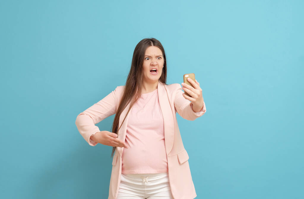 испуганная молодая беременная женщина на синем фоне звонков по мобильному телефону, выделение околоплодной жидкости, начало процесса родов - Фото, изображение