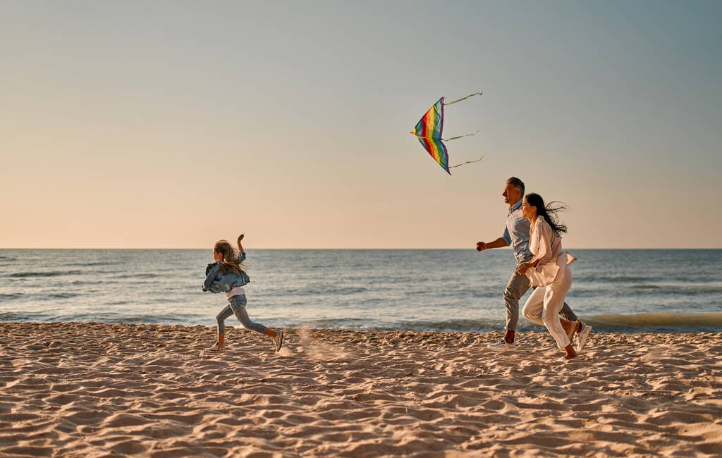 Счастливая семейная концепция. Молодая привлекательная мать, красивый отец и их маленькая милая дочь веселятся вместе на пляже. Бег с цветным воздушным змеем - Фото, изображение