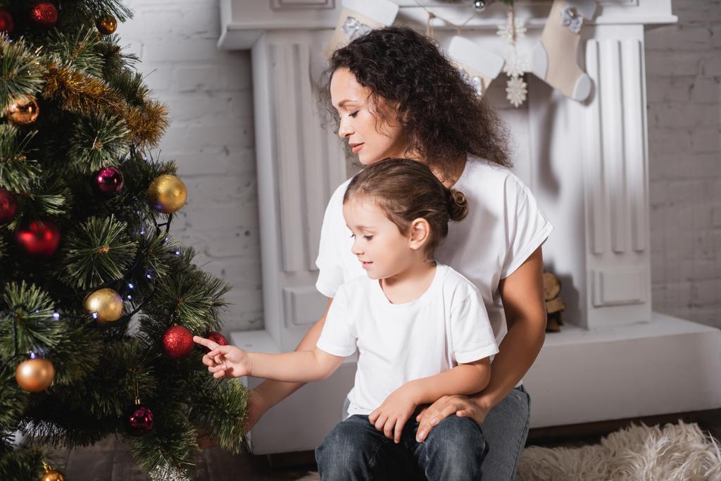 Mère et fille touchant boule de Noël sur le pin festif près de la cheminée  - Photo, image