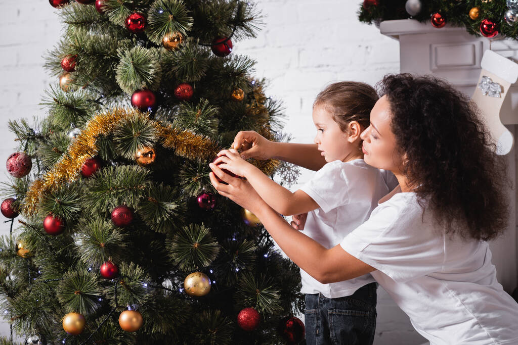 Vista laterale della mamma e della figlia che decorano il pino natalizio vicino al camino a casa - Foto, immagini