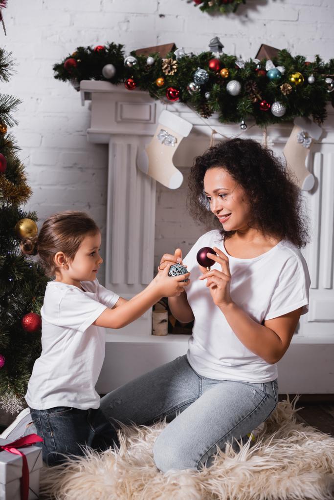 Дочь и мать с рождественскими шарами возле украшенной сосны и камина  - Фото, изображение