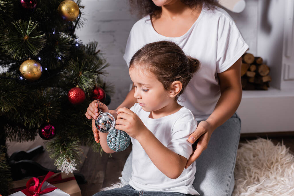 Filha sentada perto da mãe e segurando bola de Natal perto de pinho festivo em casa - Foto, Imagem