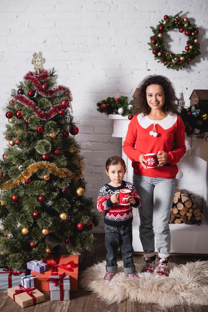 Mother and daughter with cups standing near festive pine and fireplace at home - Photo, Image