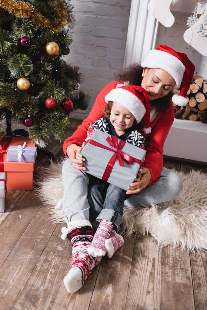 Madre abrazando hija con caja de regalo cerca de pino festivo y chimenea - Foto, imagen