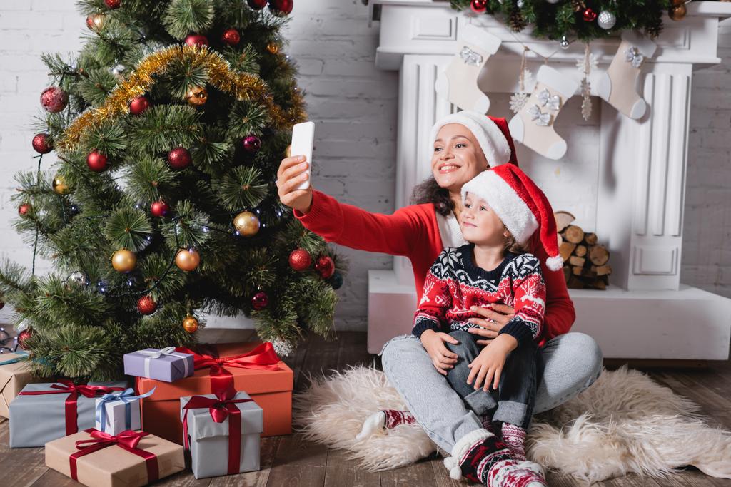 Noel Baba şapkalı anne ve kız evde çam ağacının yanında selfie çekiyorlar. - Fotoğraf, Görsel