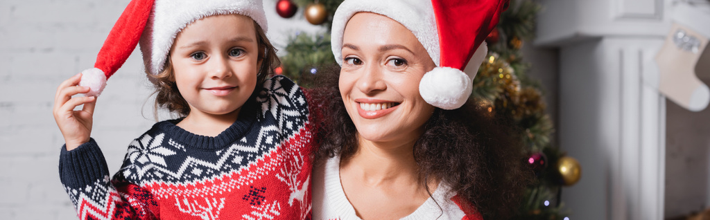 Anne ve kızının Noel Baba şapkalarıyla kameraya bakarken panoramik görüntüleri. - Fotoğraf, Görsel