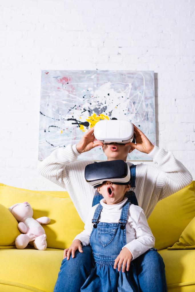 Мать и дочь сидят на жёлтом диване в наушниках виртуальной реальности - Фото, изображение