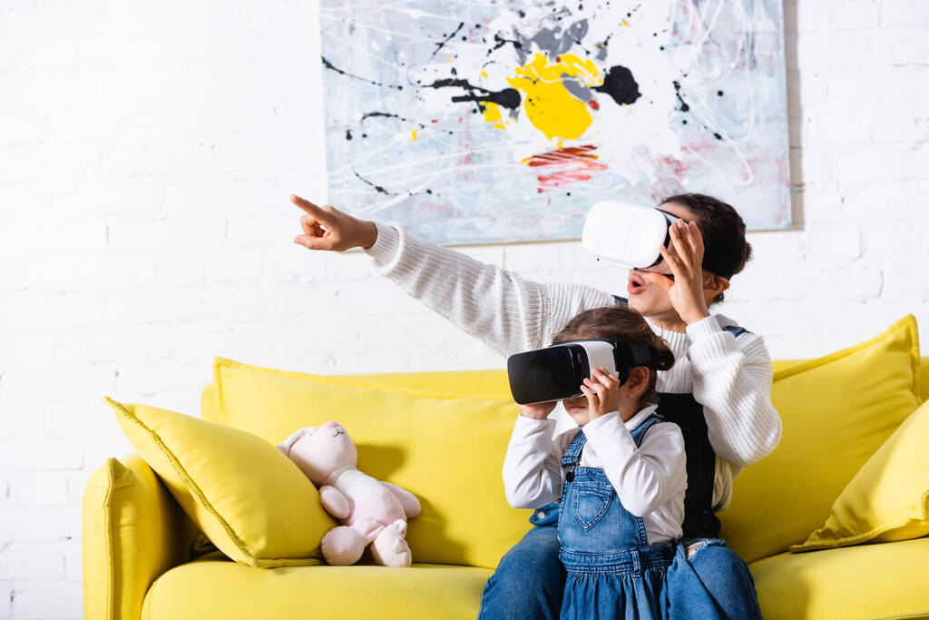 Madre señalando con el dedo y la hija usando auriculares de realidad virtual en casa - Foto, imagen