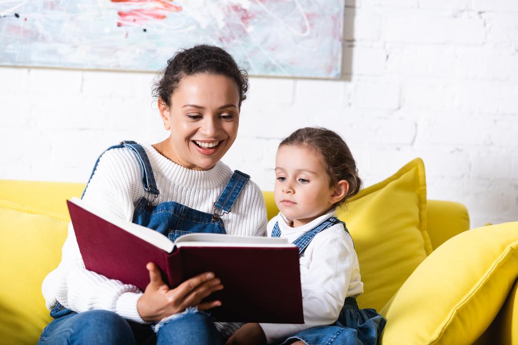 Мама и дочь читают книгу на жёлтом диване рядом с картинкой дома - Фото, изображение