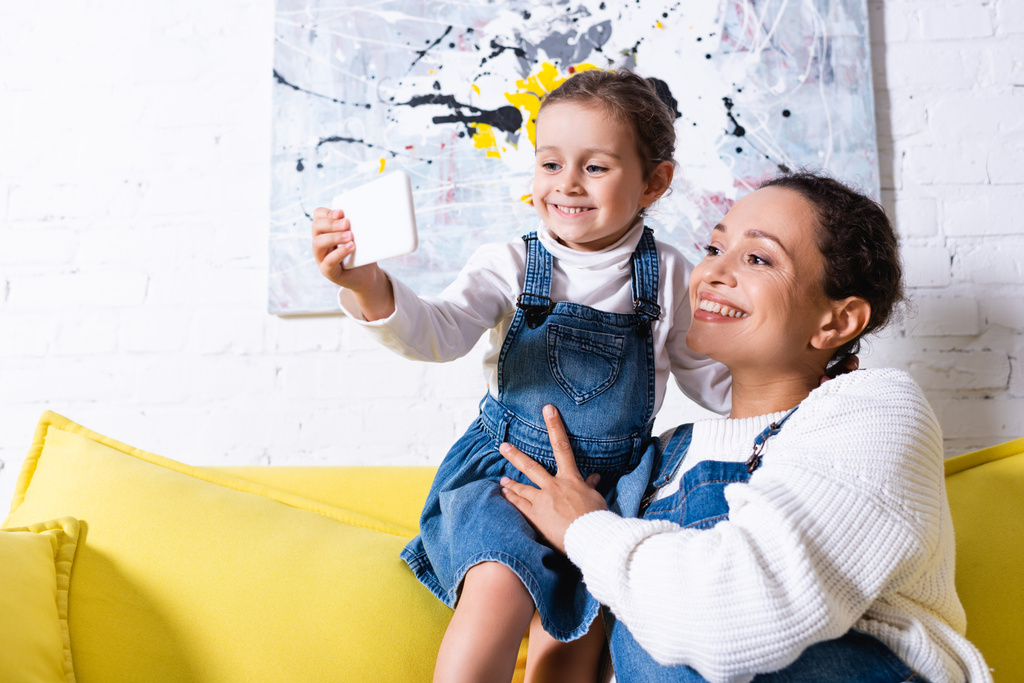 Мать и дочь делают селфи на жёлтом диване с фотографией на заднем плане - Фото, изображение