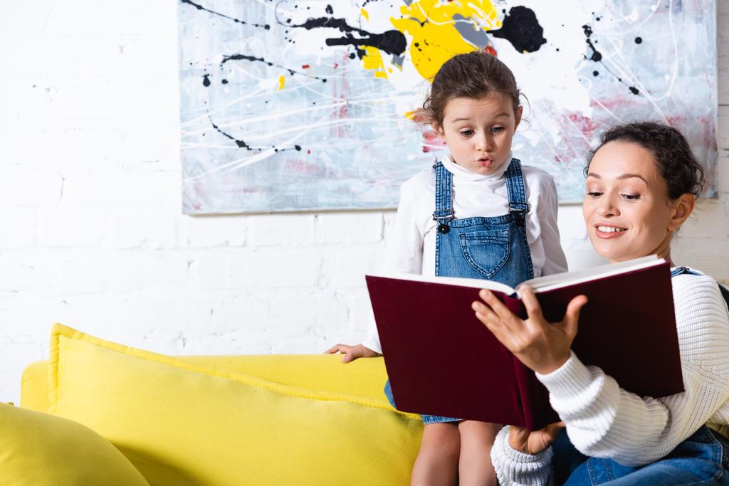 Aufgeregte Tochter steht mit Buch neben Mutter auf gelbem Sofa - Foto, Bild