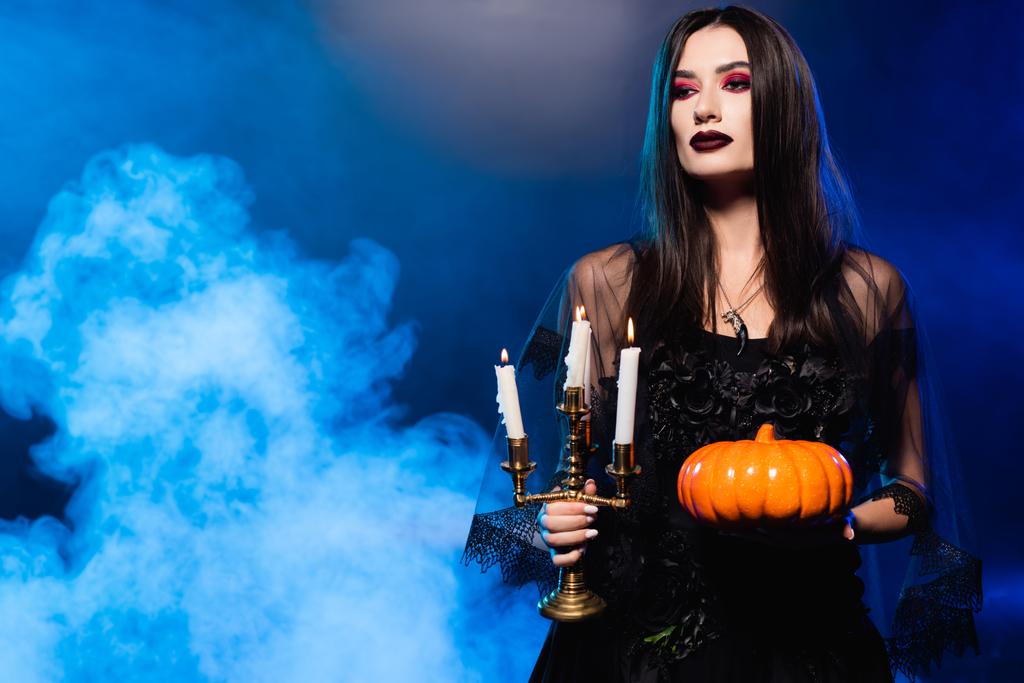 young pale woman with scary makeup holding pumpkin and burning candles on blue with smoke - Photo, Image