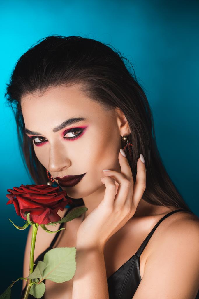 young woman with dark makeup near red rose on blue - Photo, Image