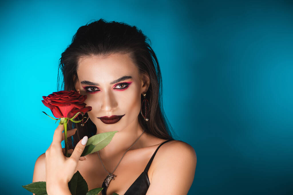 Nuori nainen tumma meikki katsomalla kameraan ja pitämällä punainen ruusu sininen - Valokuva, kuva