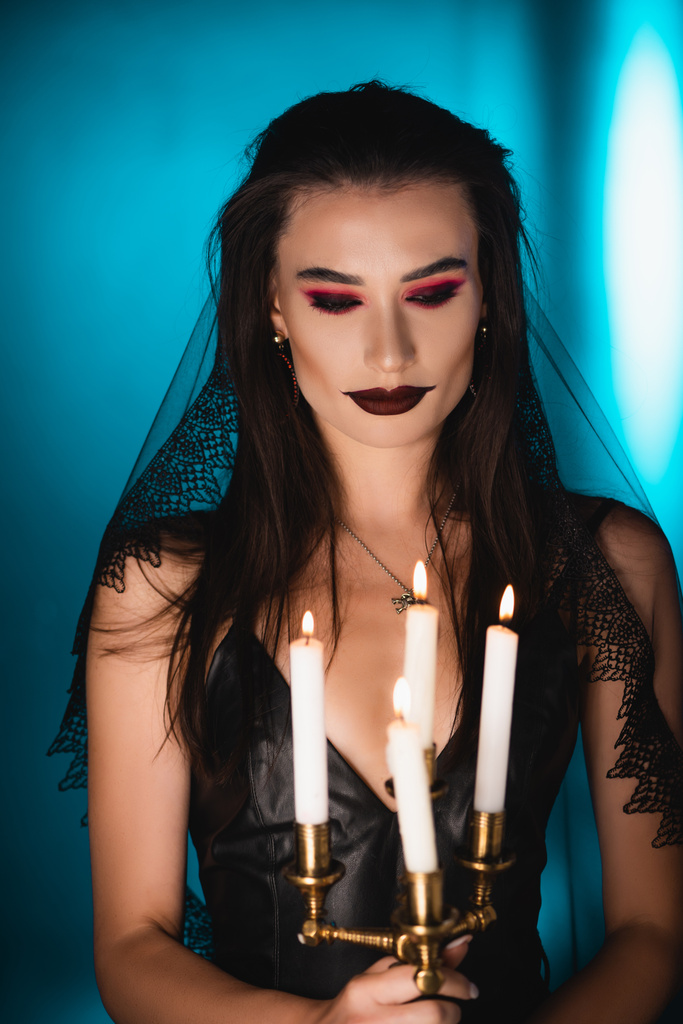 foco seletivo da mulher com maquiagem preta e véu olhando para velas acesas no azul - Foto, Imagem